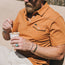 A man in a heathered cotton Ranchero Polo and sunglasses holds a cup, stirring with a spoon. He has a beard, wears rings, bracelets including a turquoise ring, and sits outdoors in the sunlight.