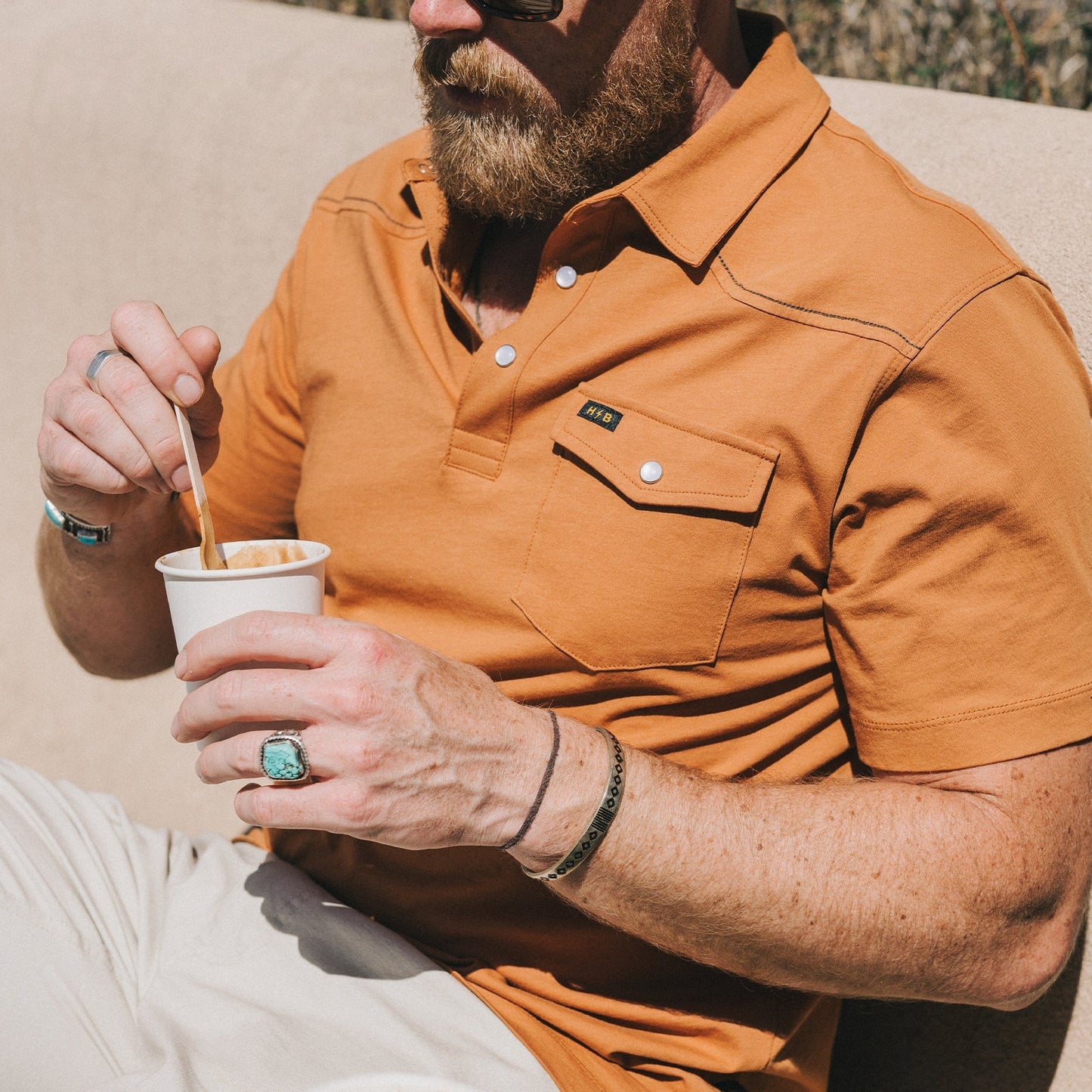 A man in a heathered cotton Ranchero Polo and sunglasses holds a cup, stirring with a spoon. He has a beard, wears rings, bracelets including a turquoise ring, and sits outdoors in the sunlight.