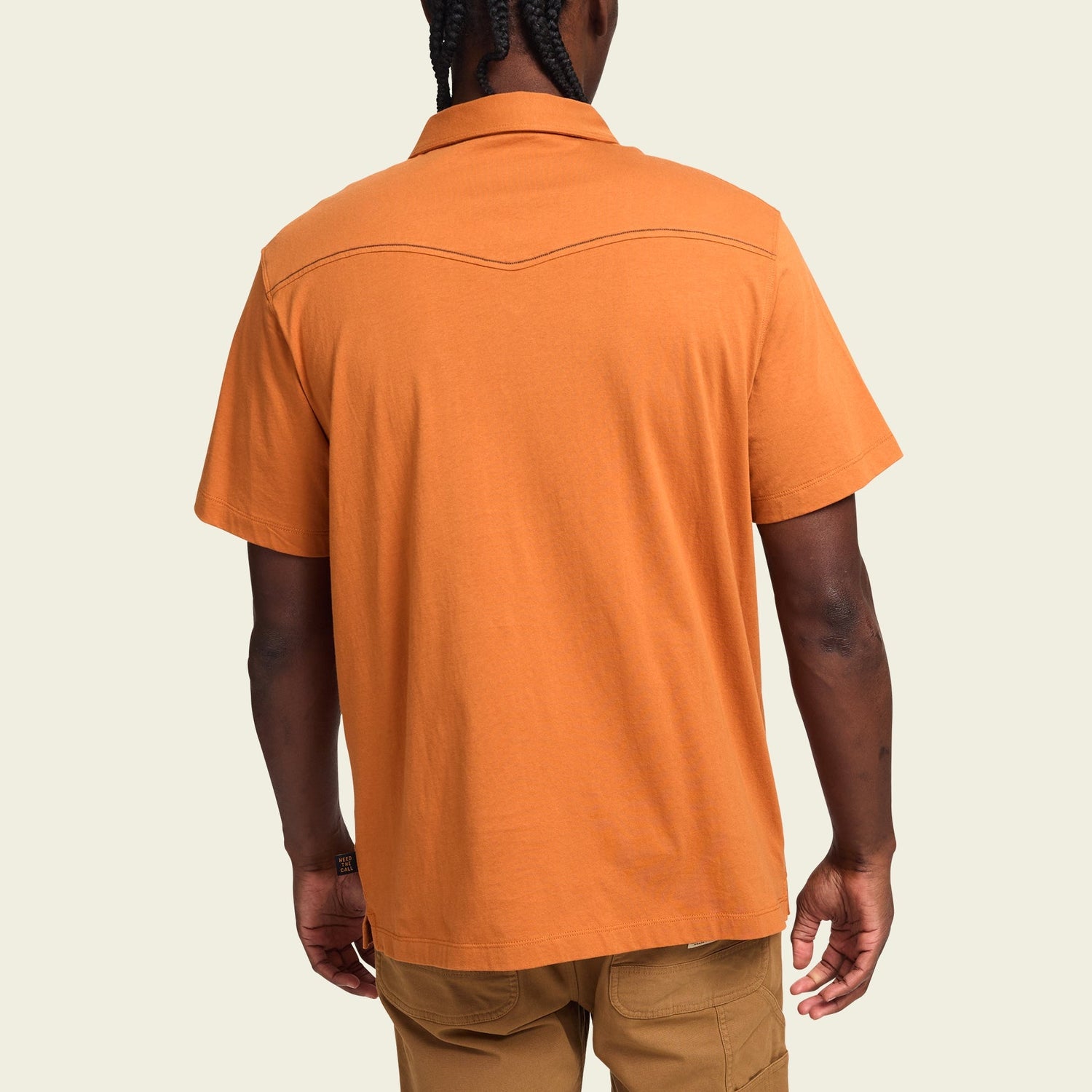 A person with braided hair is shown from the back, wearing an orange western style polo short-sleeve collared shirt crafted in heathered cotton and light brown pants, set against a plain light background.