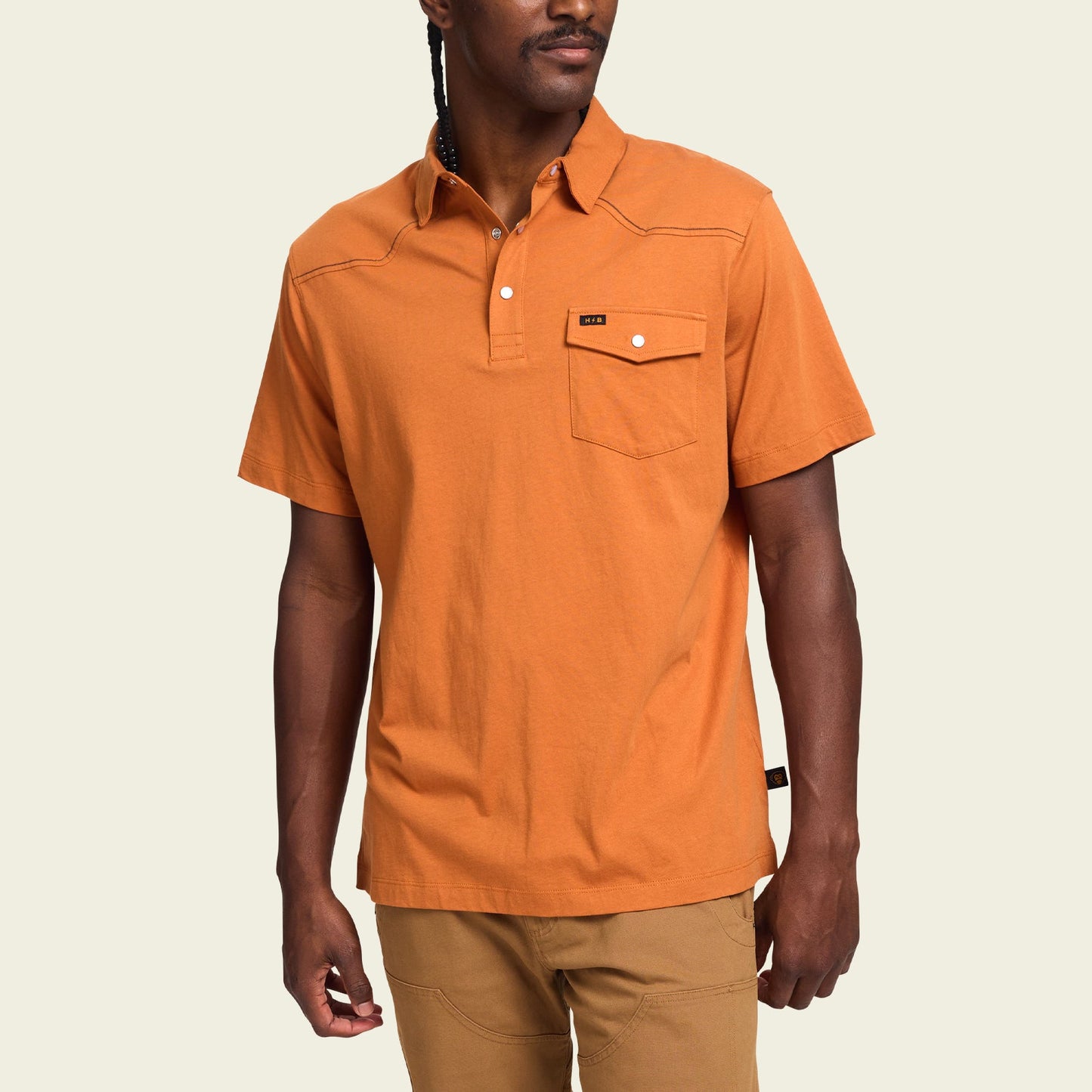 A man wearing an orange heathered cotton Ranchero Polo with a buttoned chest pocket and tan pants, standing against a plain light background.