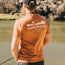 A man wearing an orange Howler blended t-shirt stands by a river, holding fishing gear, with rocky terrain and water in the background, enjoying maximum comfort from its cotton-poly blend.