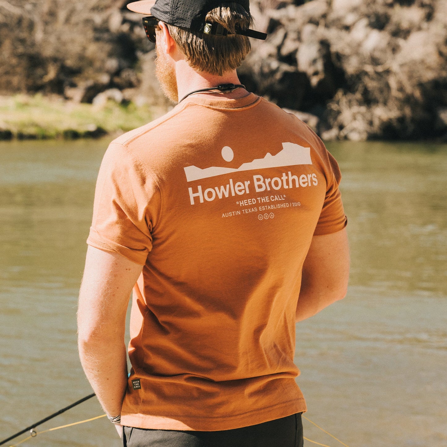 A man wearing an orange Howler blended t-shirt stands by a river, holding fishing gear, with rocky terrain and water in the background, enjoying maximum comfort from its cotton-poly blend.