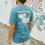 A person wearing a blue Howler blended T-shirt with a pelican graphic and text stands near a white brick wall with colorful graffiti. The cotton-poly blend ensures maximum comfort as the person faces away from the camera.