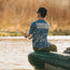 A person wearing a blue Creative Creatures Howler Brothers shirt, reminiscent of vintage t-shirts, and a cap sits on an inflatable raft while fishing on a sunlit river, with water droplets suspended in the air.