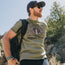 A bearded man wearing sunglasses, a cap, and an olive graphic T-shirt with HOWLER on it stands outdoors with a black backpack, evoking the laid-back style of vintage t-shirts. Trees and rocks are visible in the background.