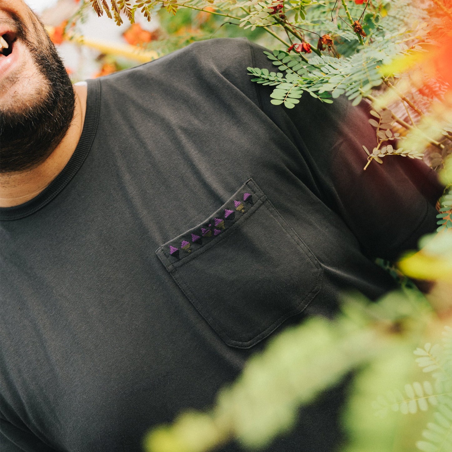 A person wearing a Howler Brothers pocket t-shirt with a patterned pocket stands among green foliage and orange flowers, smiling. Only the lower half of their face and upper torso are visible.