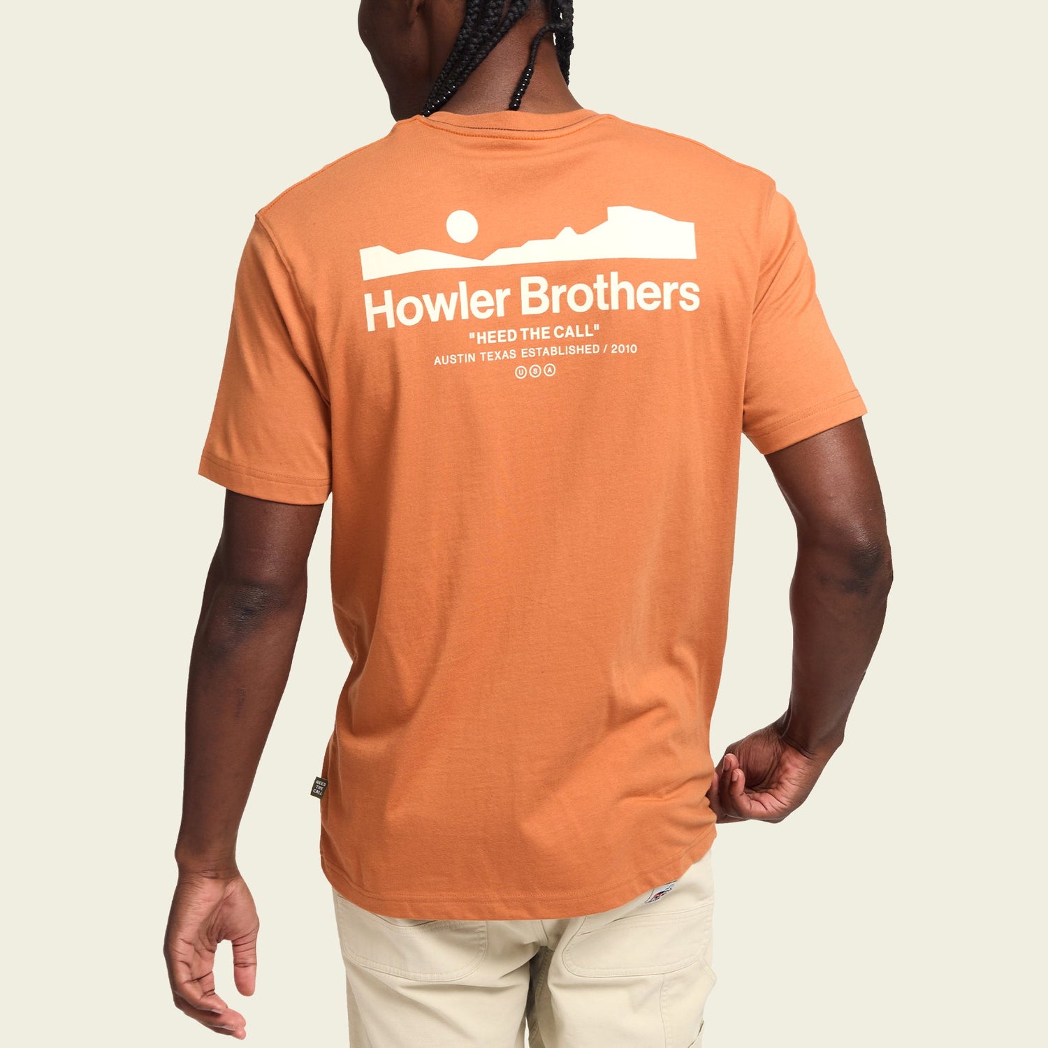 A person wearing an orange Howler blended t-shirt with white text and a landscape graphic on the back. Made from a cotton-poly blend for maximum comfort, the shirt reads “Howler Brothers ‘Heed the Call’ Austin, Texas Established 2010.” The person is facing away.