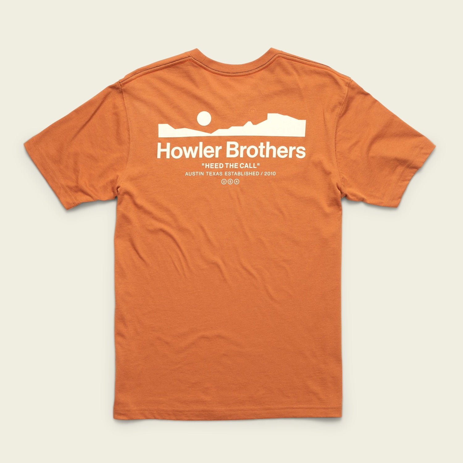 An orange cotton-poly blend t-shirt with a white graphic of a desert landscape, sun, and rock formation. The text reads Howler Brothers and Heed The Call with Austin Texas Established 2010 below for maximum comfort.