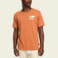 A man wearing a burnt orange Howler blended t-shirt with a small white Howler Brothers logo on the left chest, standing against a plain light background. The cotton-poly blend ensures maximum comfort, paired with light-colored pants.
