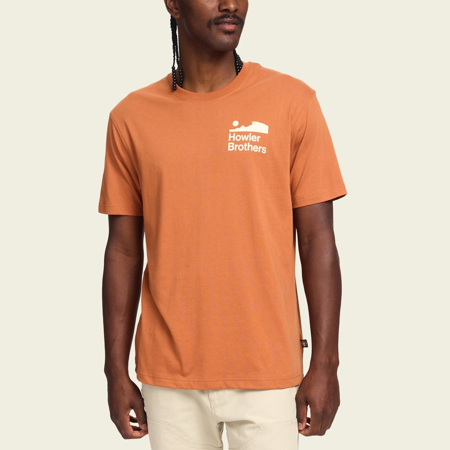 A man wearing a burnt orange Howler blended t-shirt with a small white Howler Brothers logo on the left chest, standing against a plain light background. The cotton-poly blend ensures maximum comfort, paired with light-colored pants.