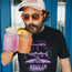 A bearded man in sunglasses and a cap drinks from two mason jar cocktails with straws and lemon garnishes, standing in front of blue double doors and wearing a black vintage t-shirt.