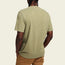A person is shown from the back wearing a light olive green short-sleeve t-shirt reminiscent of vintage t-shirts with tan pants, standing against a plain, light background.