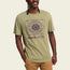 A person wearing a light olive green T-shirt with a graphic of two stylized alligators and a sun in the center, blending experimental artwork with the vibe of vintage t-shirts. The text reads “HOWLER BROTHER.” The person smiles, one hand in their pocket.