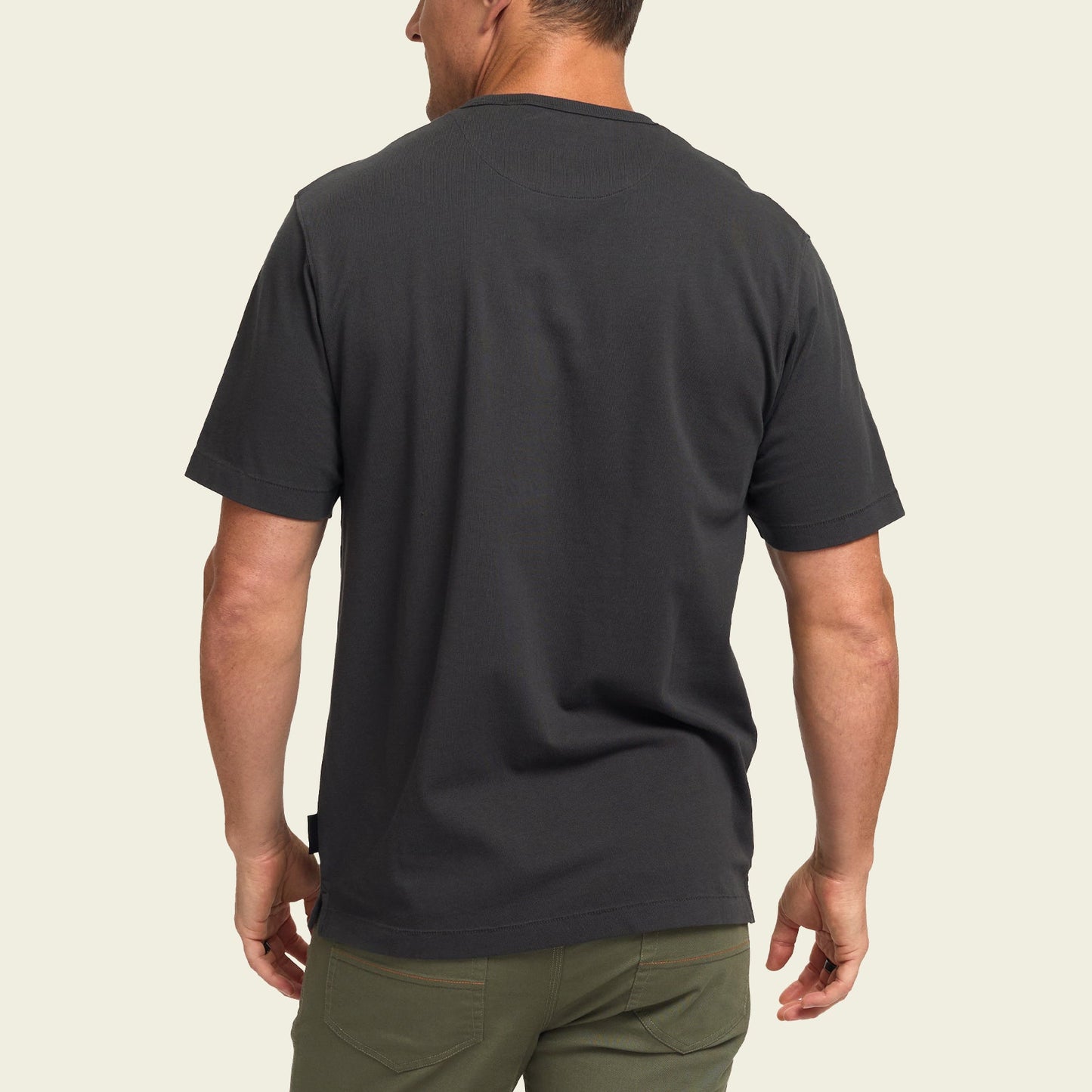 A man is shown from the back wearing a plain black heavyweight cotton t-shirt and olive green pants, standing against a light beige background.