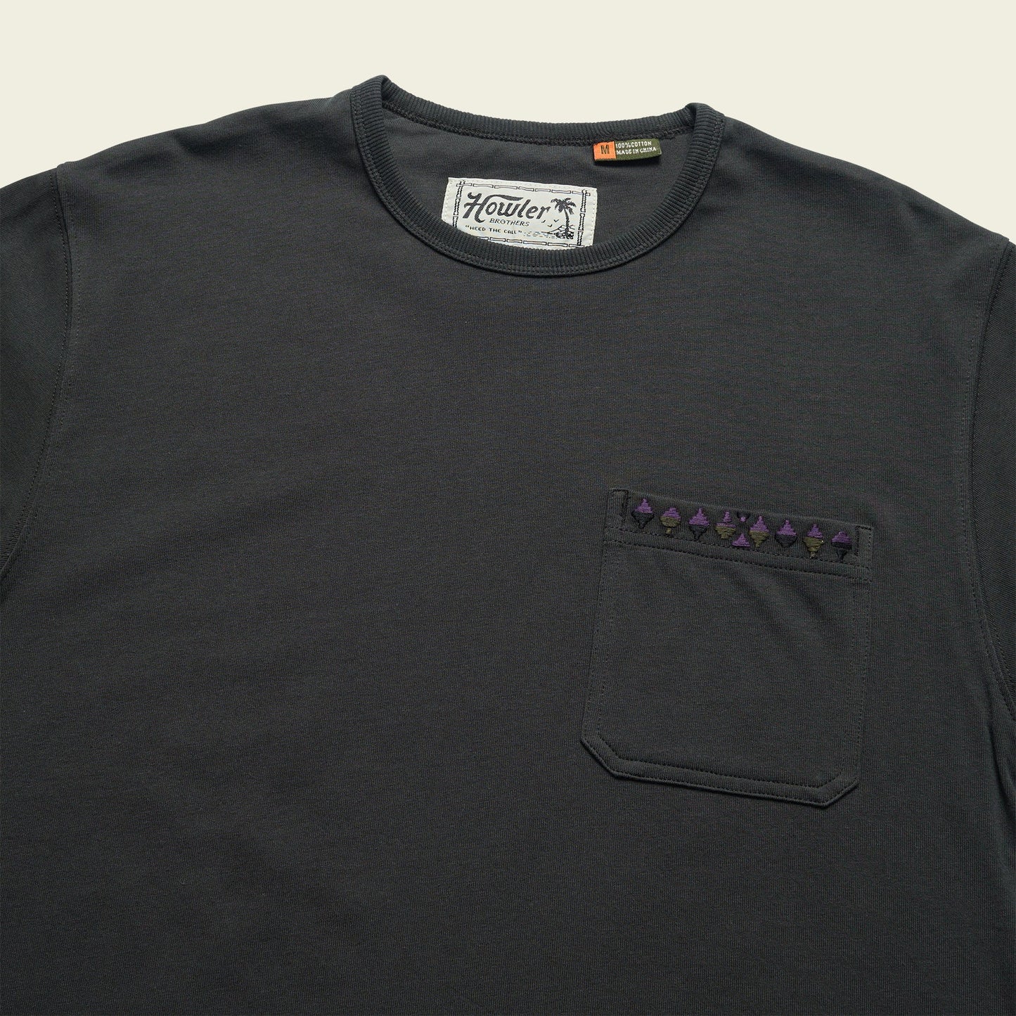 A dark gray Howler Brothers pocket t-shirt made from heavyweight cotton, featuring a chest pocket with a row of small purple geometric shapes along the top edge. The label inside the collar reads Howler.