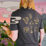 A person with curly blonde hair, seen from behind, wears a comfortable Howler t-shirt with gold mystical designs and holds a chalice. Illustrated smoke and sparkles overlay a vibrant outdoor scene of mountains, flowers, and a river.
