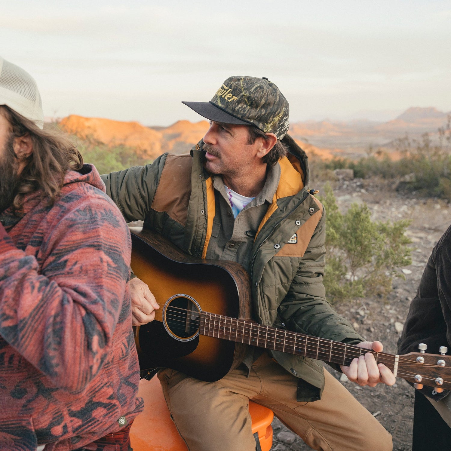 A man in a jacket and camo soft crown hat with an adjustable snapback closure plays an acoustic guitar outdoors in a desert landscape, sitting beside another person in a patterned hoodie. Mountains and scrubby plants stretch across the background.