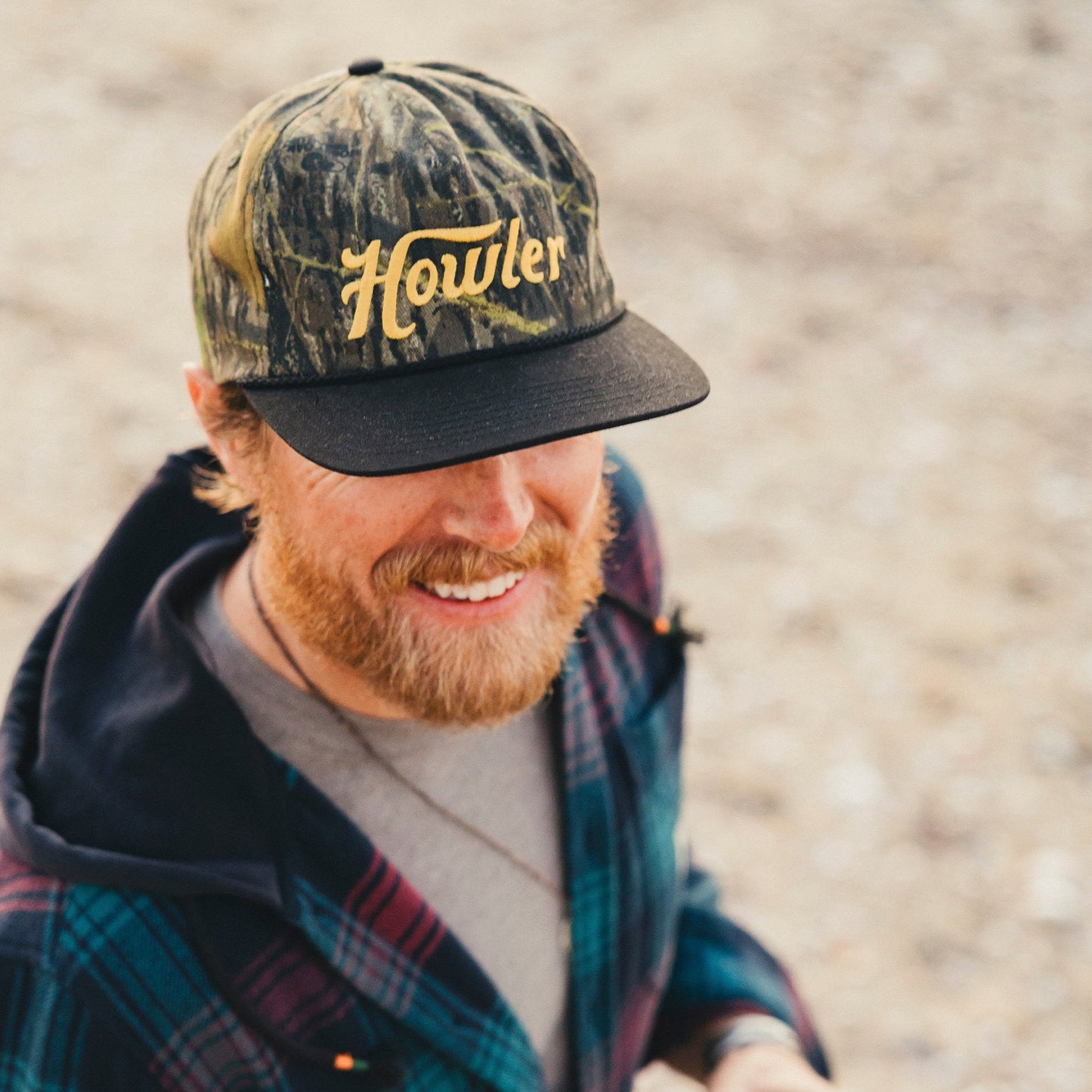A smiling man with a reddish beard wears a camouflage Howler cap featuring a soft crown and adjustable snapback closure, along with a plaid jacket, standing outdoors on a blurred, sandy background.