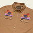A tan Crosscut Deluxe button-up shirt with vintage inspired embroidery above the front pockets, depicting colorful desert landscapes with mountains, trees, and sunset clouds. The shirt label reads Howler.