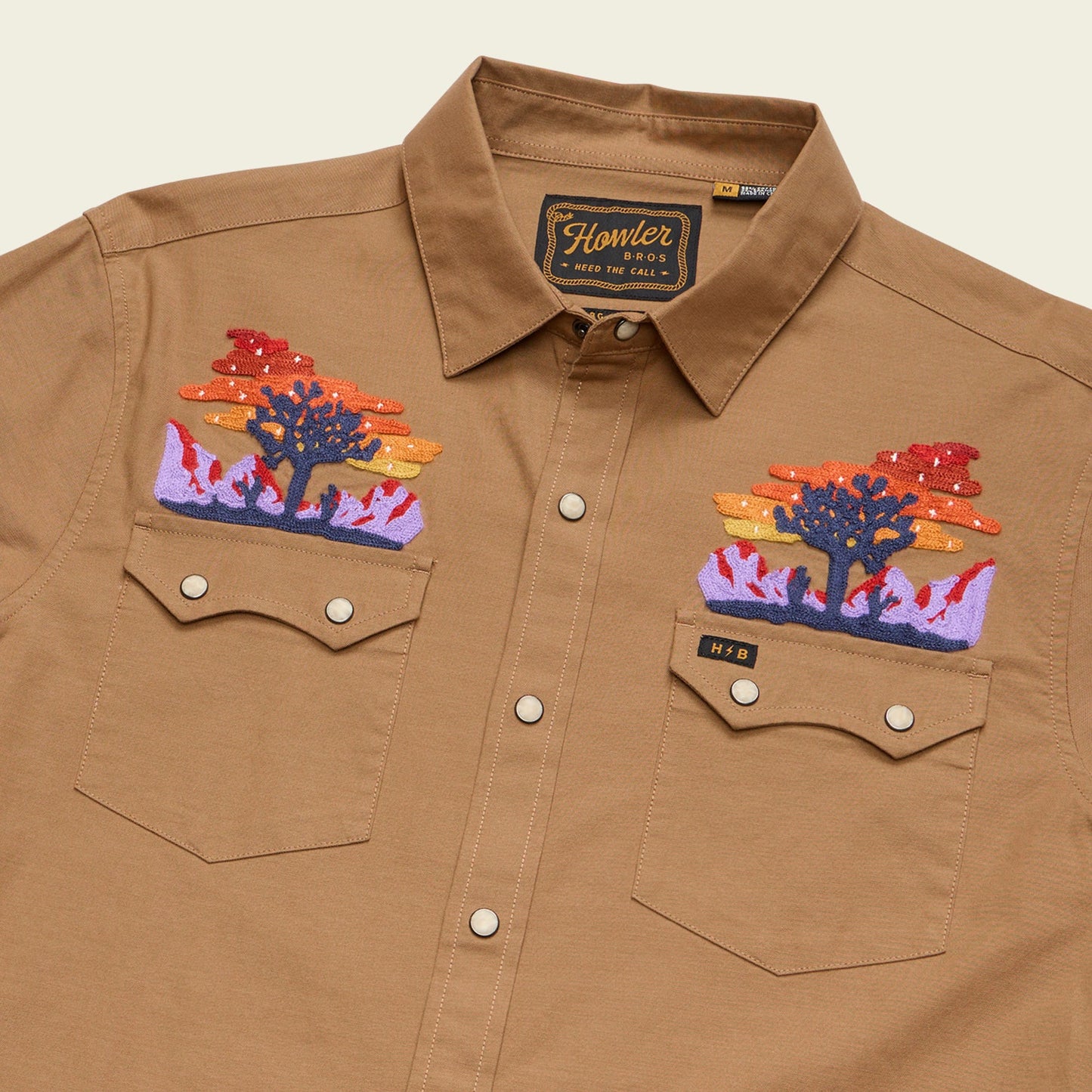 A tan Crosscut Deluxe button-up shirt with vintage inspired embroidery above the front pockets, depicting colorful desert landscapes with mountains, trees, and sunset clouds. The shirt label reads Howler.