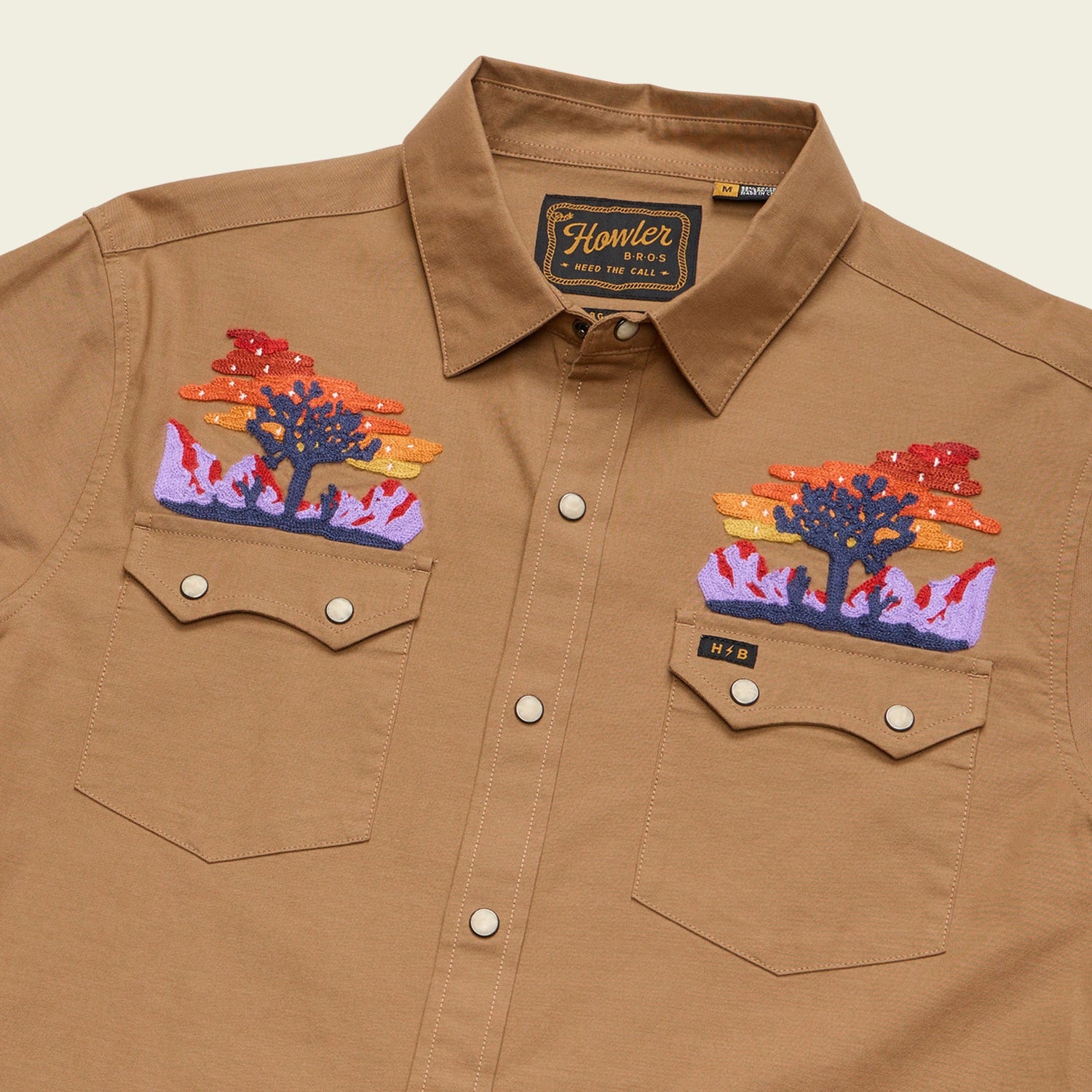 A tan Crosscut Deluxe button-up shirt with vintage inspired embroidery above the front pockets, depicting colorful desert landscapes with mountains, trees, and sunset clouds. The shirt label reads Howler.