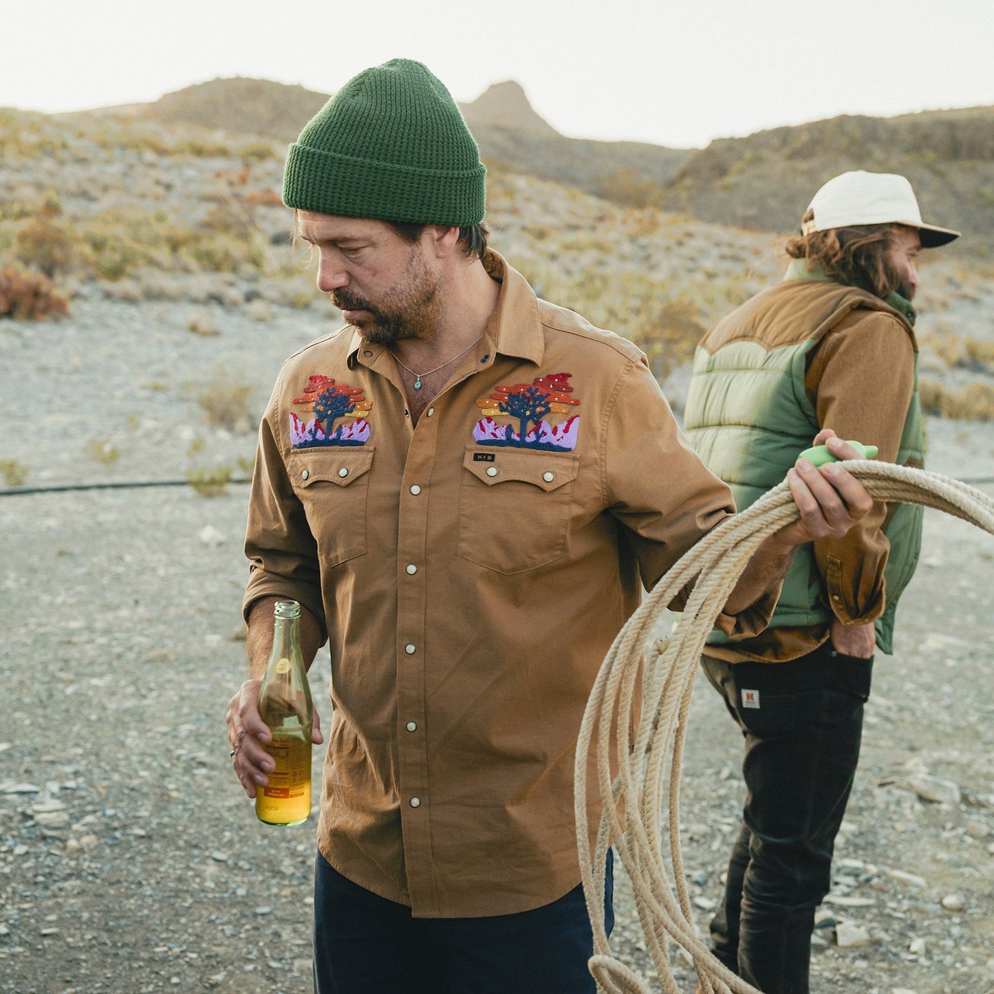 A man in a brown shirt featuring vintage inspired embroidery and a green beanie holds a beer and a rope outdoors, while another man in a green vest and hat stands in the background amid a rocky, open landscape.
