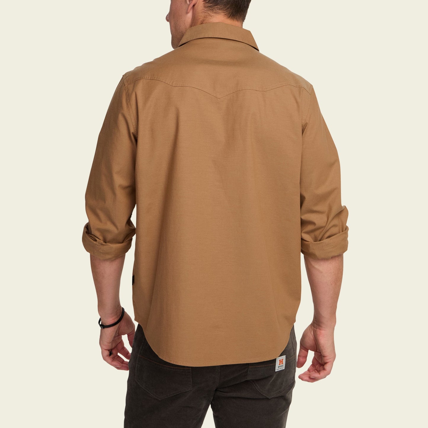 A man stands with his back to the camera, wearing a light brown Crosscut Deluxe long-sleeve shirt with rolled-up sleeves and black pants, featuring vintage inspired embroidery against a plain, light background.