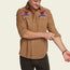 A man in a tan stretch chambray button-up with vintage inspired embroidery on the chest rolls up his sleeve. He is smiling and looking down, standing against a plain light background.