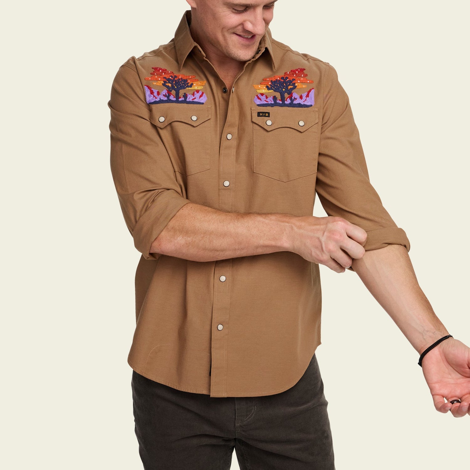 A man in a tan stretch chambray button-up with vintage inspired embroidery on the chest rolls up his sleeve. He is smiling and looking down, standing against a plain light background.