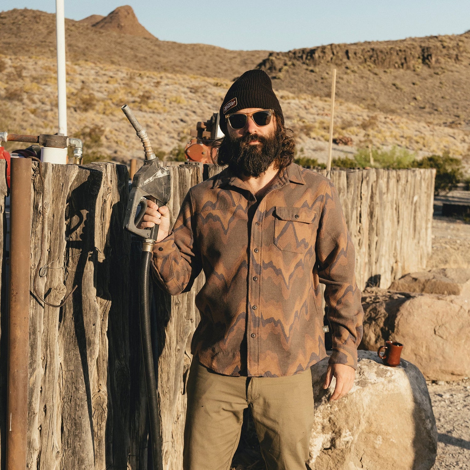 A bearded man wearing sunglasses, a beanie, and a buttery soft flannel stands outdoors by a rustic wooden fence, holding a metal kettle in one hand. Desert mountains and dry vegetation are visible in the background.