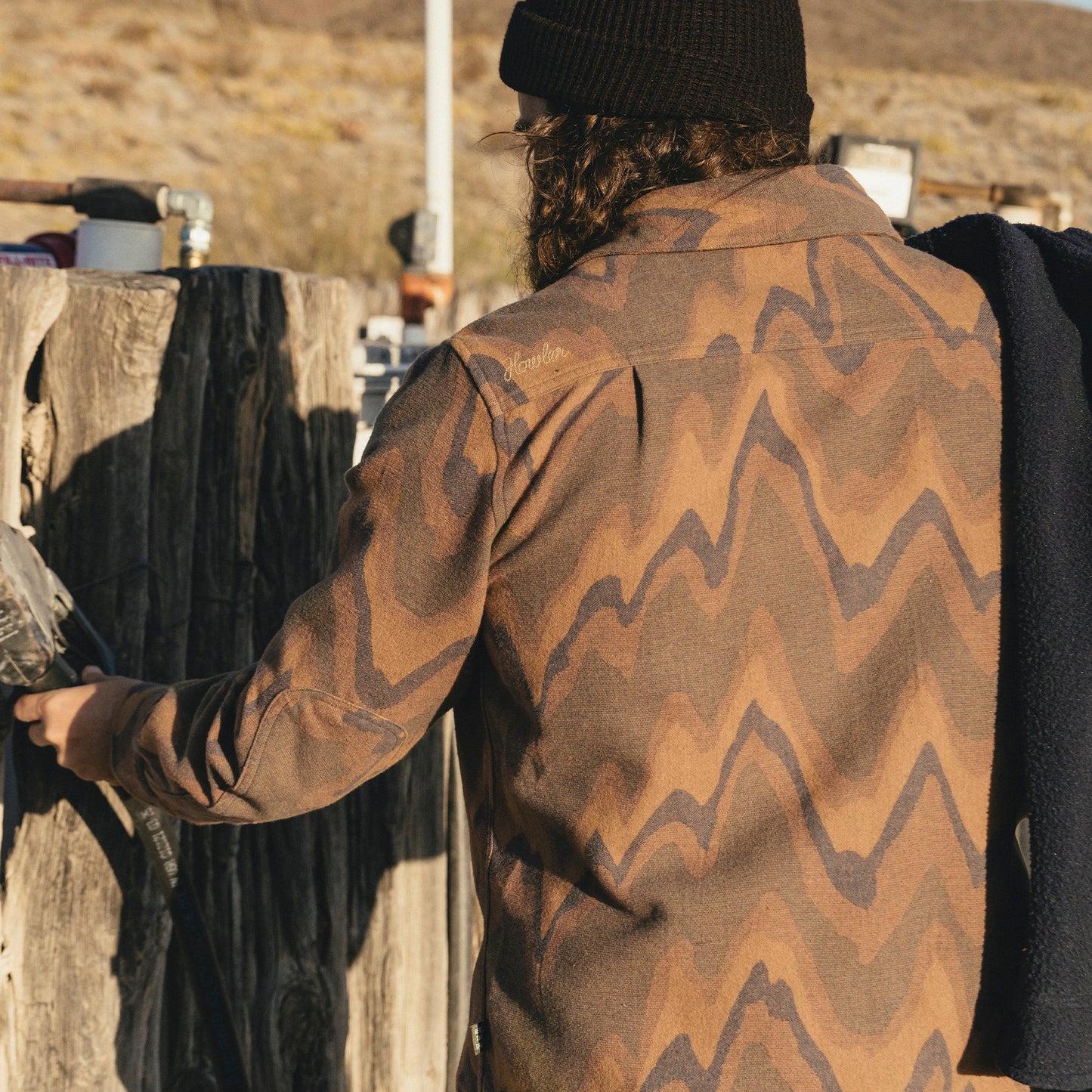 A person with long hair, wearing a black beanie and a brown zigzag-patterned Harker’s Flannel jacket, stands outdoors by a wooden fence, holding a paintbrush and a navy garment. Sunlight casts warm tones on the buttery soft flannel.