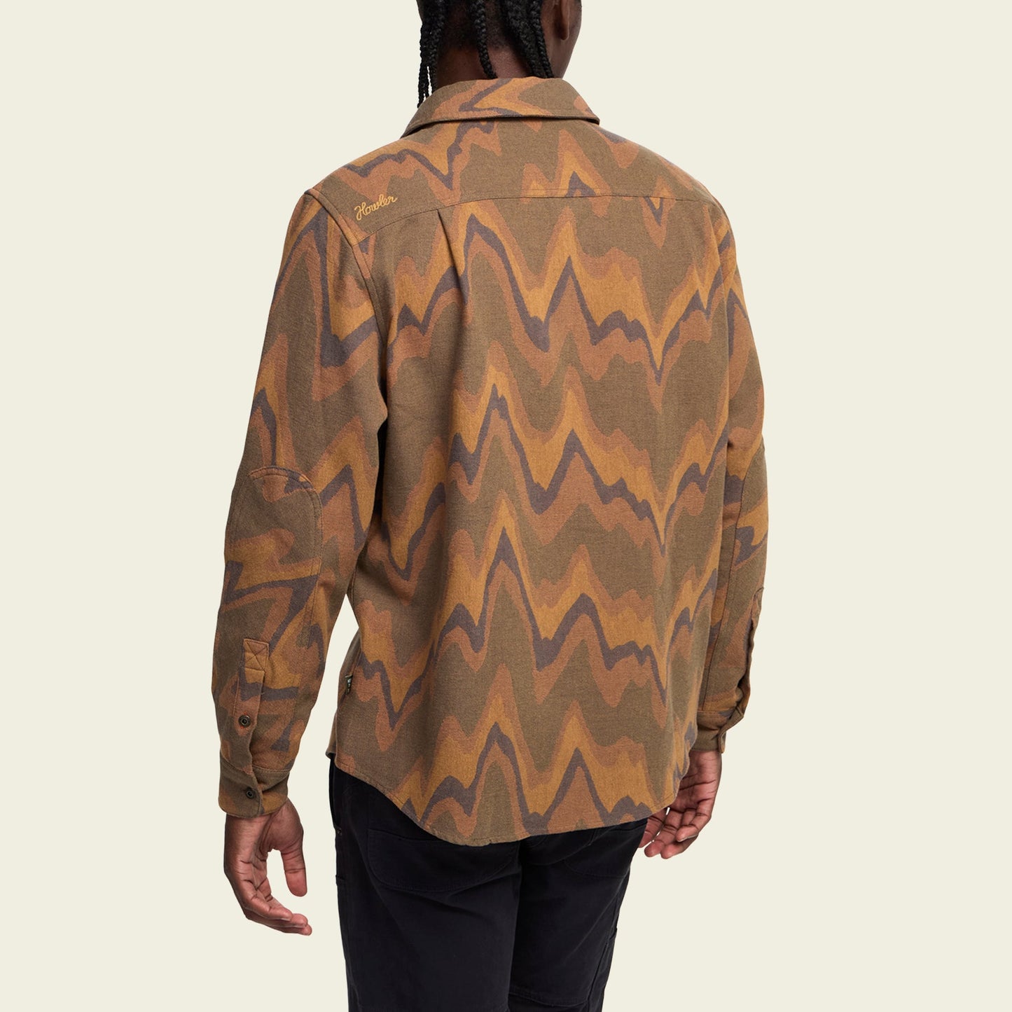 A person with braided hair is shown from the back, wearing a buttery soft flannel long-sleeve shirt with a brown, orange, and tan wavy pattern and black pants against a plain light background.