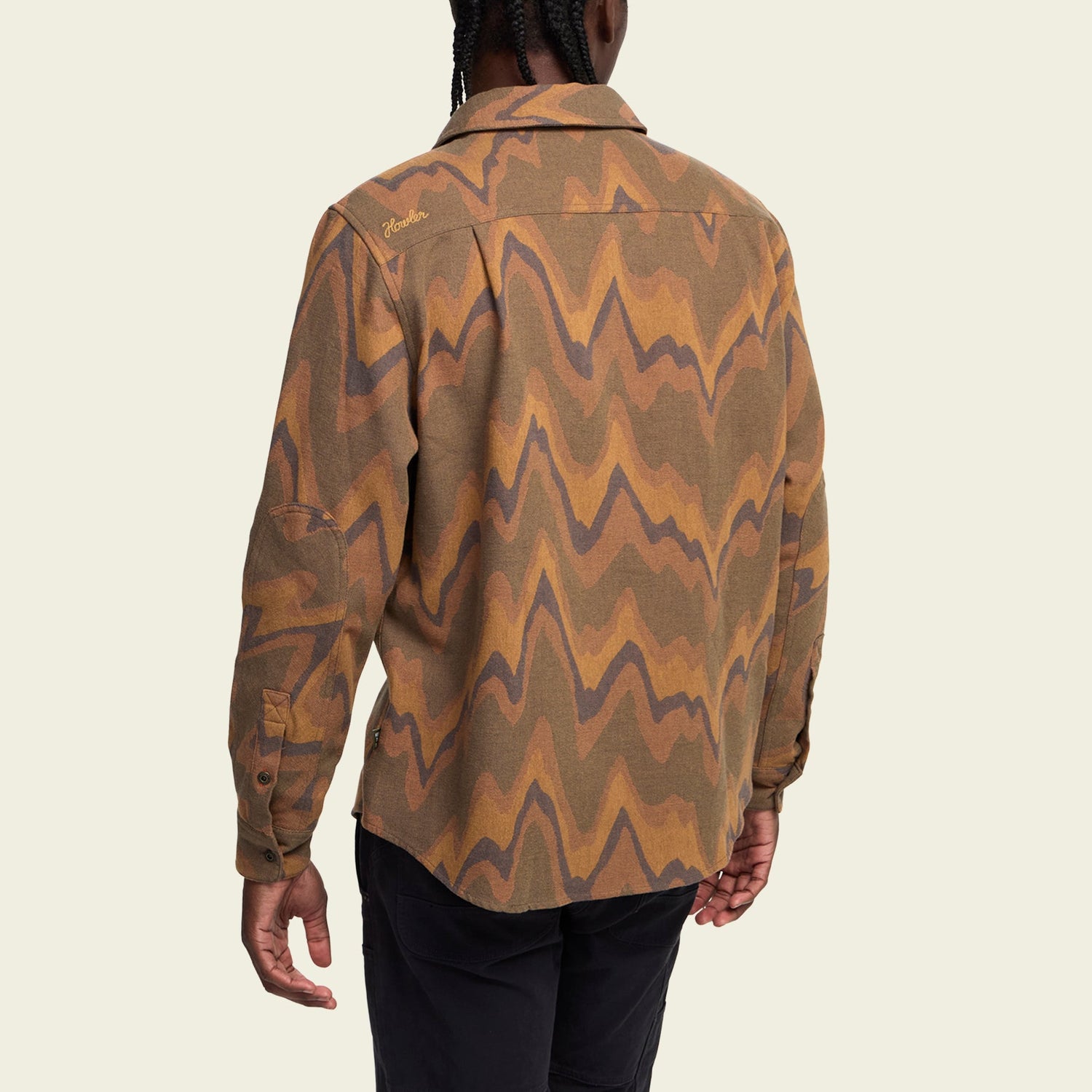 A person with braided hair is shown from the back, wearing a buttery soft flannel long-sleeve shirt with a brown, orange, and tan wavy pattern and black pants against a plain light background.