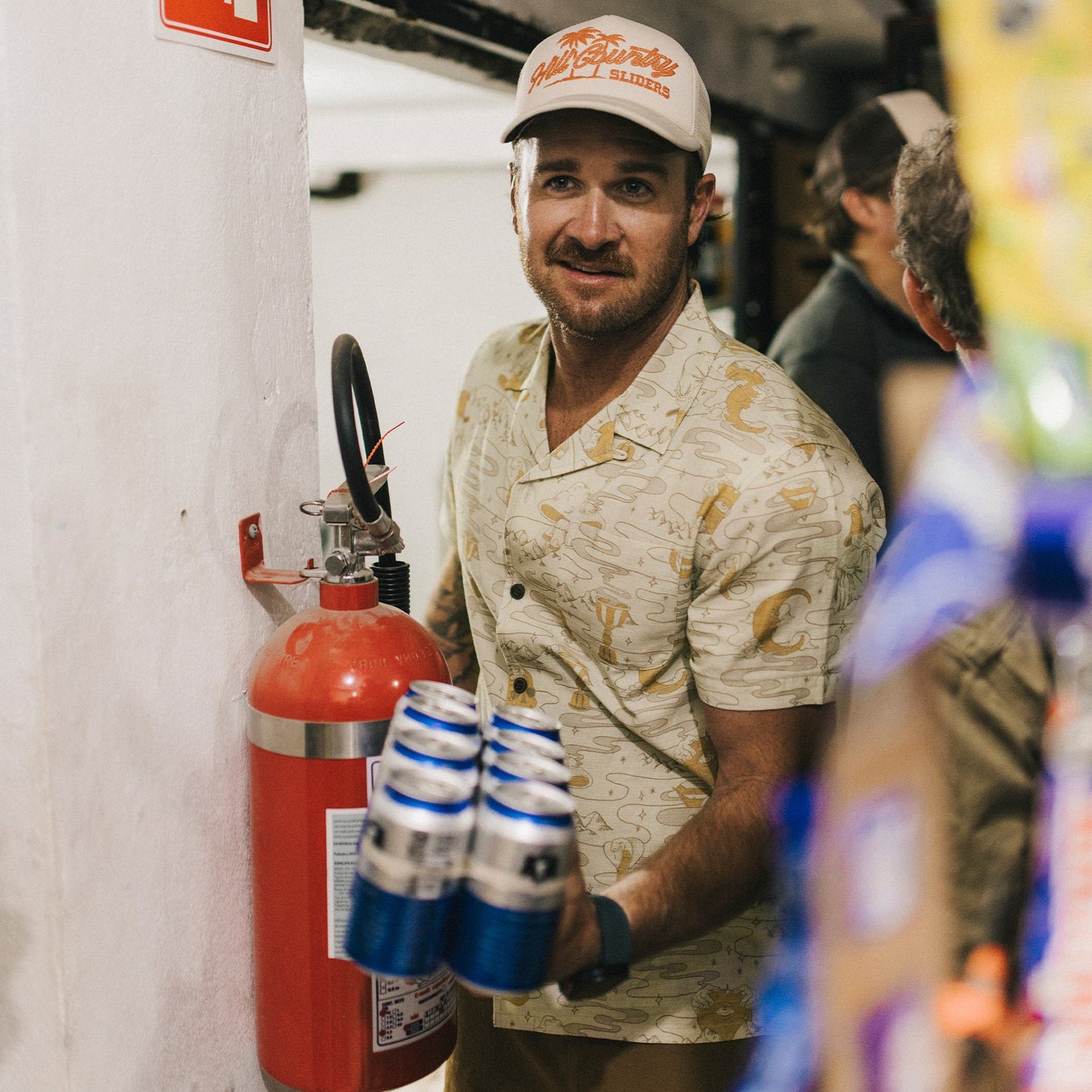 A man in a vintage Hawaiian shirt with a camp collar stands by a fire extinguisher, holding several cans of beer in one hand and looking toward the camera in an indoor setting.