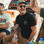 Three friends sit on the tailgate of a truck, laughing and enjoying drinks. One in a black cotton t-shirt and green cap smiles widely, while another in a tie-dye shirt with a vintage look grins beside him.