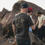 Three men stand outdoors by large rocks. The man in the foreground wears a black Howler t-shirt with yellow illustrations and a black cap. Two other men, one in a red cap and another in a beige shirt, explore the rocks.