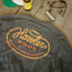 A dark Howler blended t-shirt with “Howler Bros, Austin, TX, Welcome to Howler Country” in orange text lies on a wooden surface next to a beer bottle, sunglasses, dice, a harmonica, and the brim of a green and white cap.