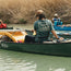 A person wearing a green Howler blended t-shirt and a tan cap paddles a green canoe on a river, with another person and canoe visible near tall grasses, enjoying maximum comfort and adventure.