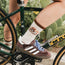 A person wearing Adidas sneakers and white socks with a floral design from the Flower Power Collection stands next to a green bicycle, with cactus plants in the background.
