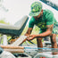 A man in a green shirt and camouflage snapback inspects equipment on a boat, surrounded by greenery. He is leaning forward, focused on his task, with supplies and gear visible around him.