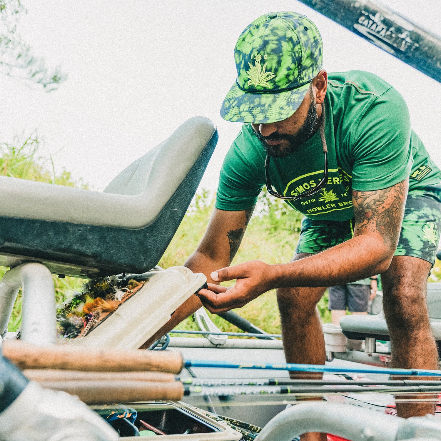 A man in a green shirt and camouflage snapback inspects equipment on a boat, surrounded by greenery. He is leaning forward, focused on his task, with supplies and gear visible around him.