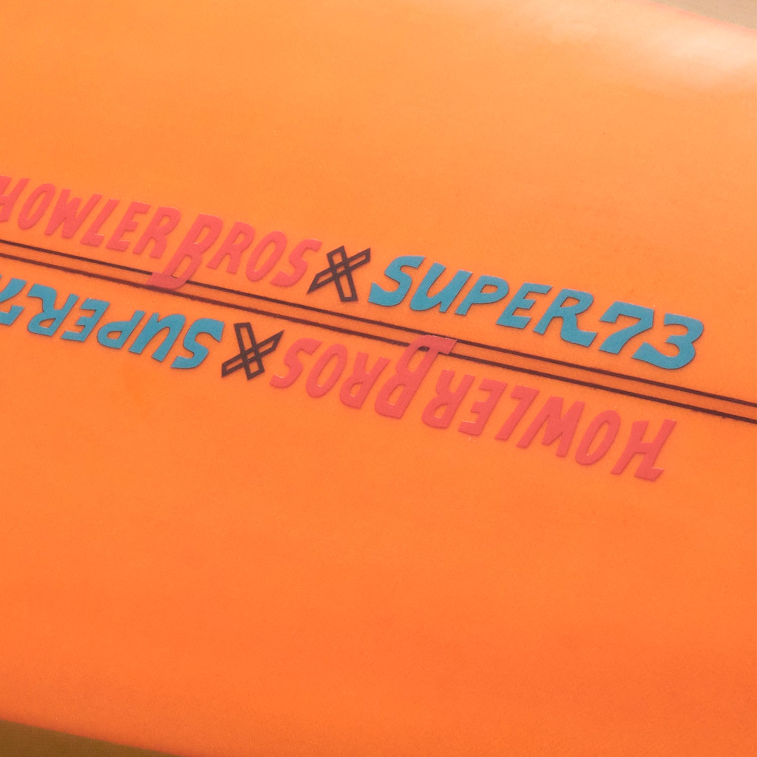 Close-up of orange surface featuring HOWLER BROS x SUPER73 repeated in pink and blue text, separated by a black line—highlighting the vibe of a Lightning Powered Surf Machine.