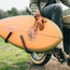 A person wearing brown leather gloves holds an Almond Surfboards orange surfboard while sitting on a Super73-S2 bicycle with surfboard racks, on a grassy outdoor path.