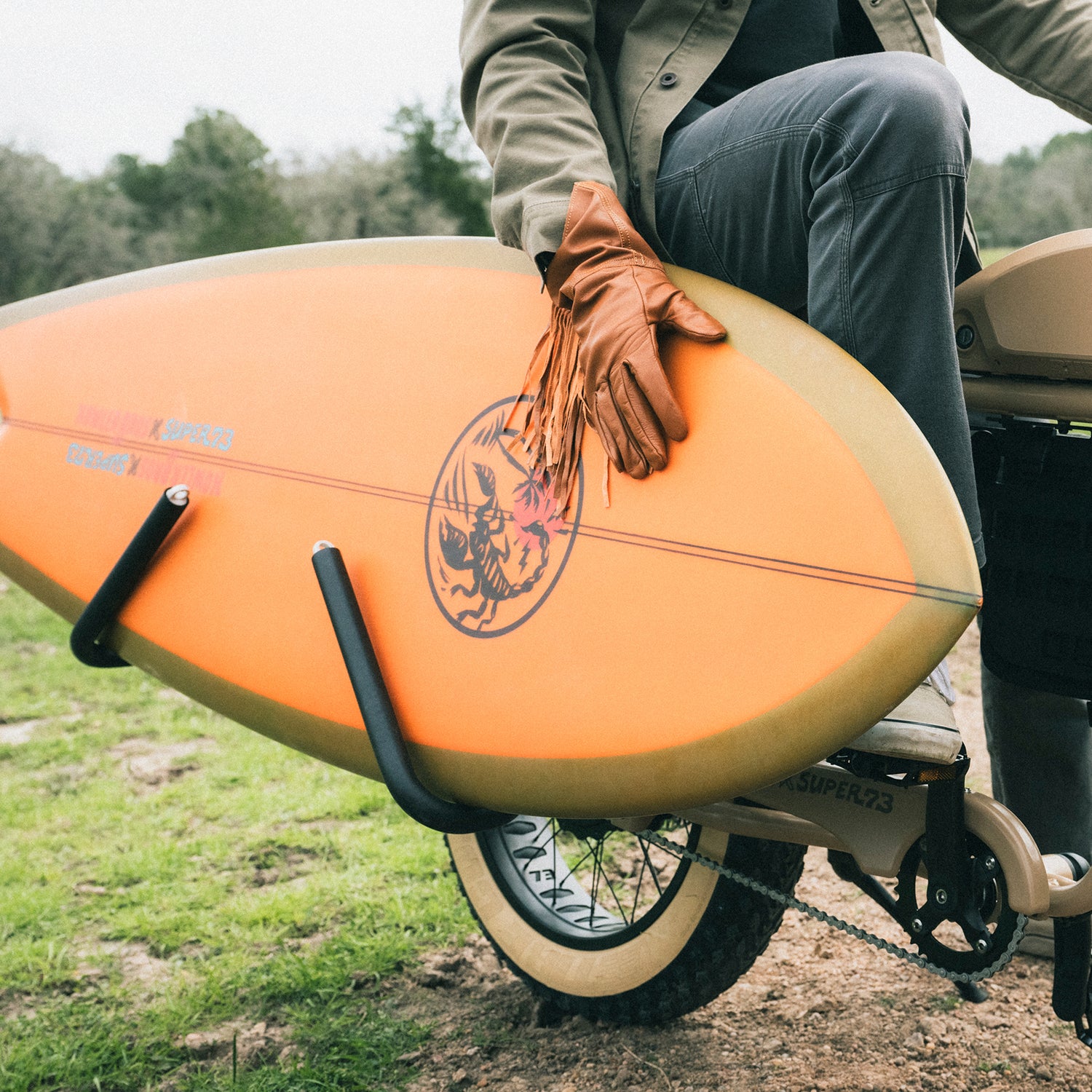 A person wearing brown leather gloves holds an Almond Surfboards orange surfboard while sitting on a Super73-S2 bicycle with surfboard racks, on a grassy outdoor path.