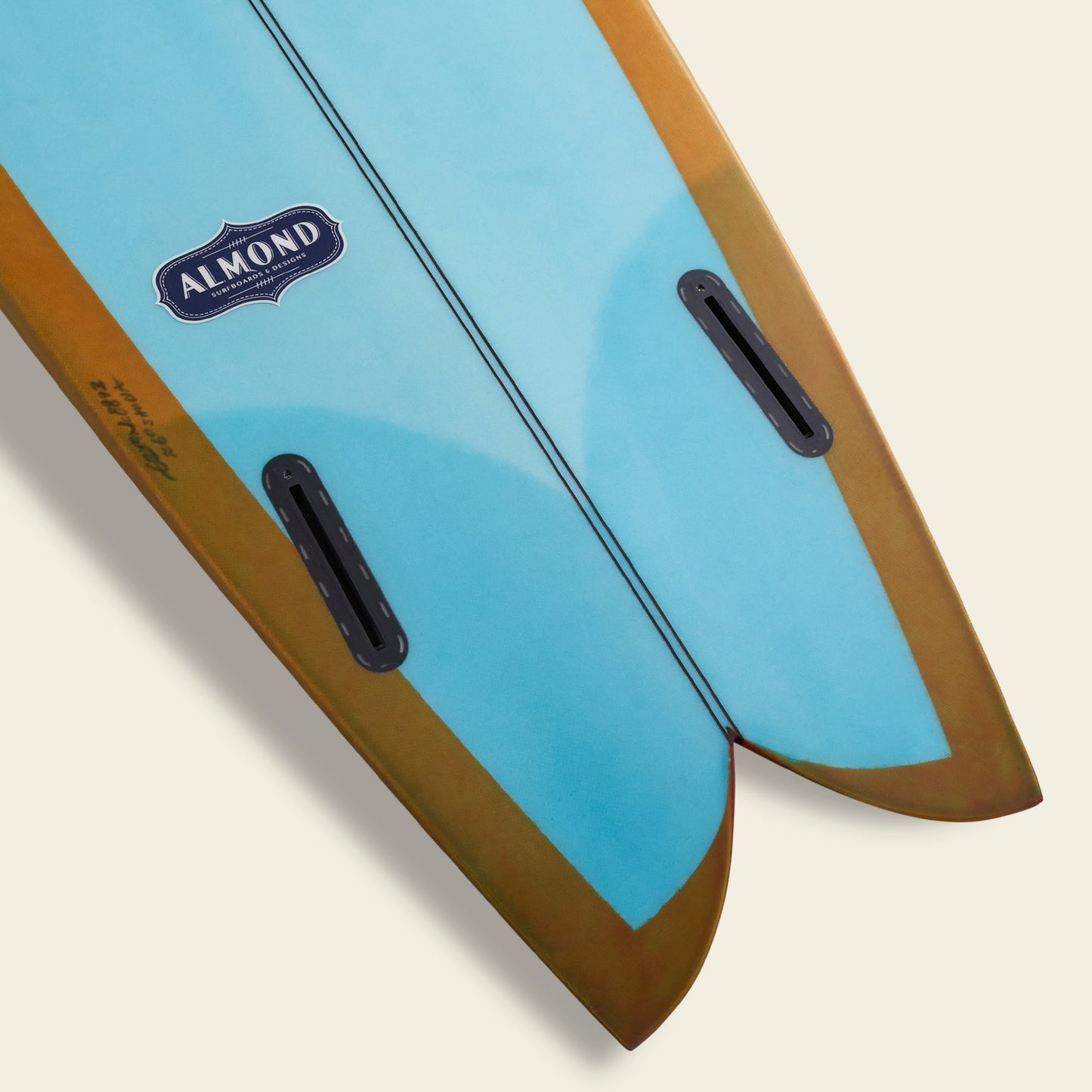 Close-up of a blue and brown twin-fin surfboard by Almond Surfboards, featuring the ALMOND logo near the tail, two fin boxes per side, and a split tail design—showcasing a true Lightning Powered Surf Machine against a light background.
