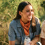 A young woman with long brown hair, wearing a blue shirt and a Los Longhorns de Tejas Bandana, sits outdoors smiling and holding a can. Sunlight and greenery are in the background, perfect for showing off her game day accessories.