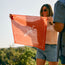 A woman in sunglasses and denim shorts smiles outdoors, holding up a Los Longhorns de Tejas Bandana—one of the must-have game day accessories—against a backdrop of green trees on a sunny day.