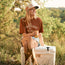 A woman in a cap, Howler blended t-shirt, denim shorts, and cowboy boots sits on a white cooler outdoors, smiling and holding a can. Green foliage and sunlight fill the background.