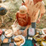 A woman in a University of Texas Longhorns shirt and Foam Dome hat prepares food at an outdoor picnic table filled with bread, cheese, packaged food, and burgers, while a man stands beside her holding grilling tools. A grill smokes in the background.