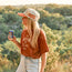 A woman in a red T-shirt and denim shorts stands outdoors, holding a can of soda. She sports sunglasses and a vintage-inspired hat, with greenery and hills bathed in warm sunlight as her scenic backdrop.